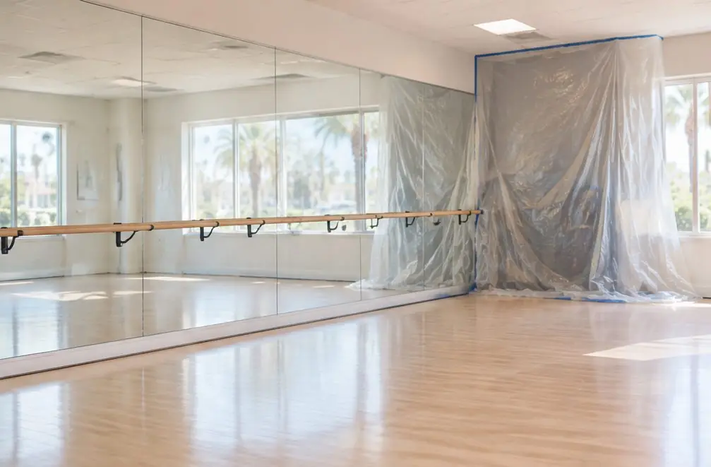 Illustration: a private dance studio in a Chula Vista, CA business park with a pale maple floor, a wall of mirrors, a ballet barre and plastic sheeting covering the corner